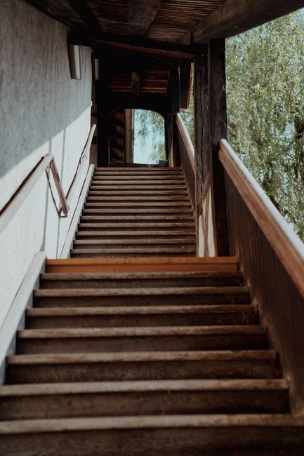 A narrow wooden staircase with evenly spaced steps, viewed from the bottom looking upward, inside a residential property. The staircase features a wooden handrail on the right side and is enclosed by