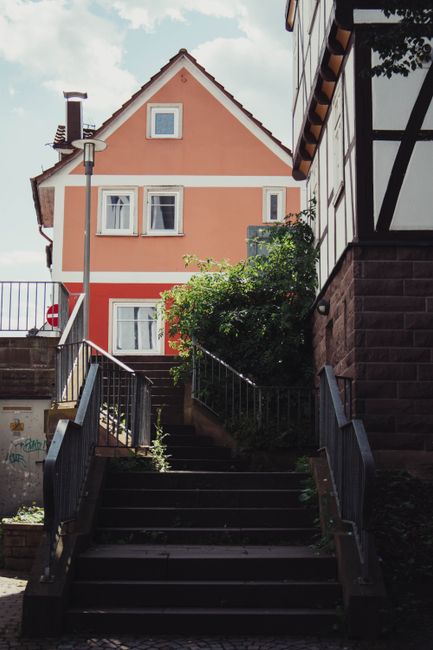 A narrow wooden staircase with evenly spaced steps, viewed from the bottom looking upward, inside a residential property. The staircase features a wooden handrail on the right side and is enclosed by